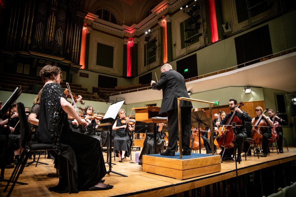 Hibernian Orchestra performing live on stage with conductor John Finucane, string players, and full orchestral ensemble in a concert hall