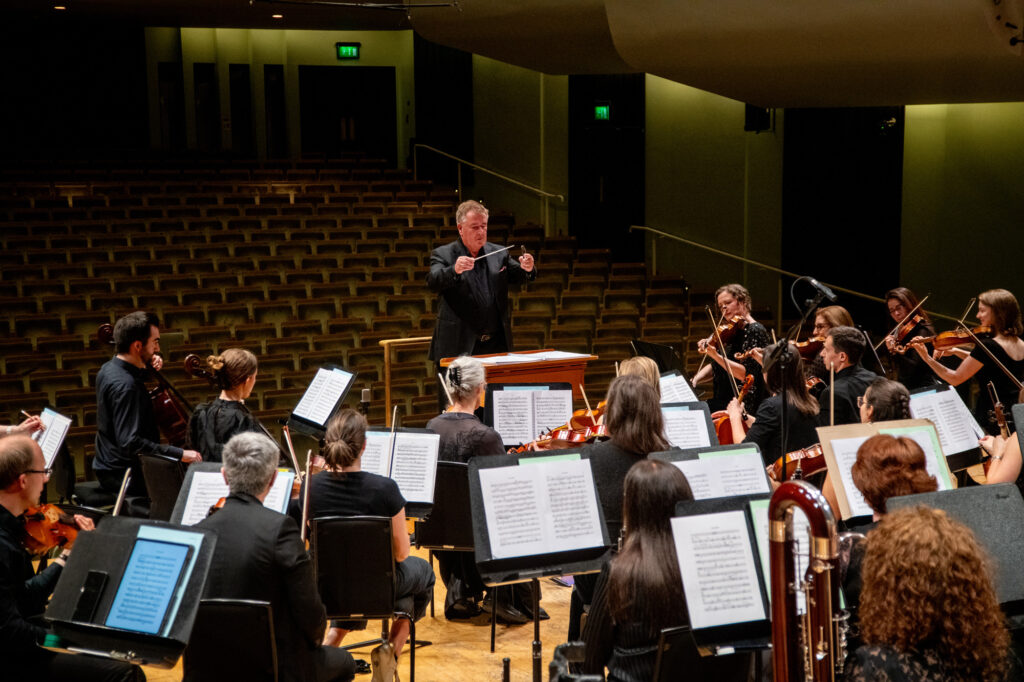 Hibernian Orchestra conductor leading a rehearsal with full string section and music stands visible in the concert hall