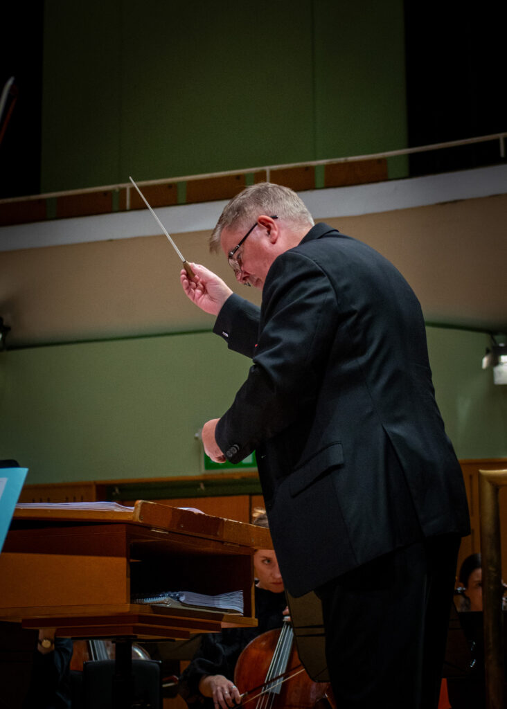 Hibernian Orchestra conductor John Finucane leading an orchestral rehearsal in close-up on the podium with musicians visible in the background