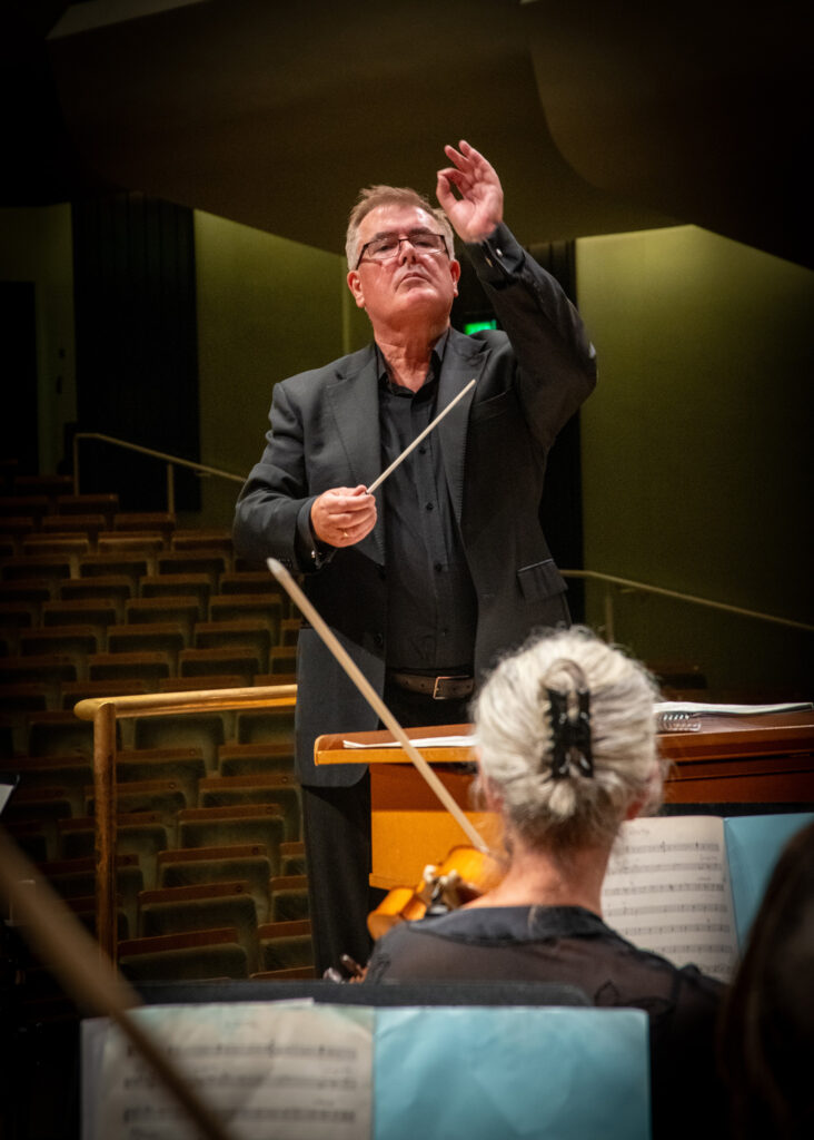 Hibernian Orchestra conductor in close-up leading rehearsal with baton raised and orchestra musicians visible in the foreground
