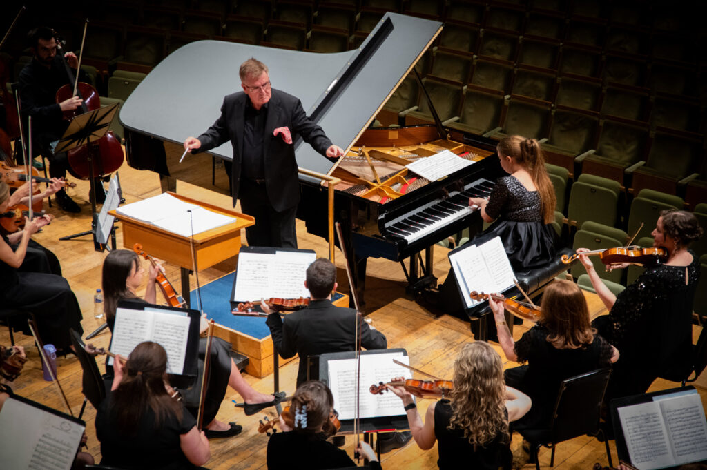 Hibernian Orchestra rehearsal with conductor on podium, piano, and string players performing in the concert hall