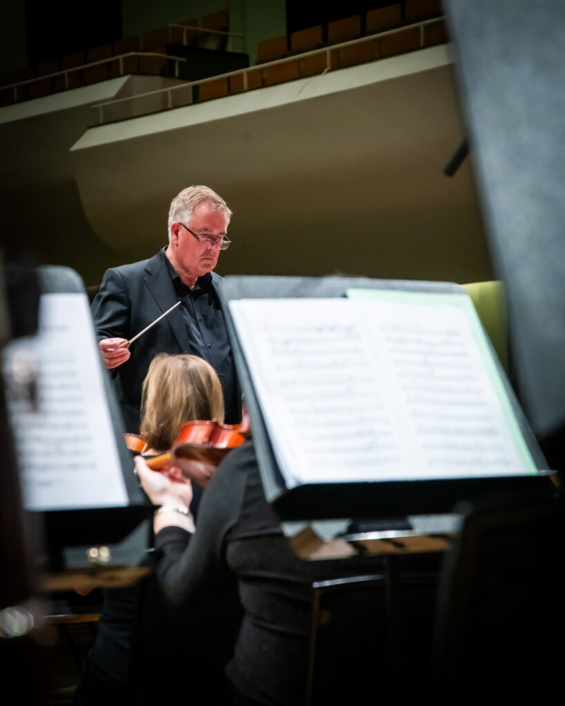 Hibernian Orchestra conductor leading rehearsal with music stands blurred in the foreground and orchestra musicians seated on stage